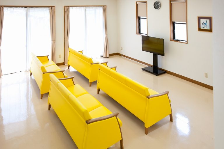 黄色いソファが配置された明るい共用リビングスペース。大きな窓と壁掛けテレビがある。